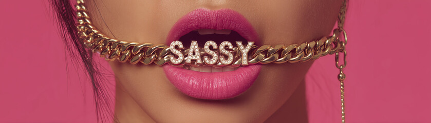 closeup of woman holding up gold chain necklace in her mouth with big pink lips and the gold necklace says "SASSY" in diamonds, edgy fashion jewelry design statement