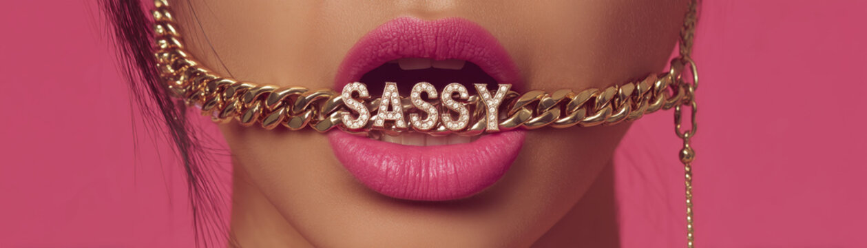 closeup of woman holding up gold chain necklace in her mouth with big pink lips and the gold necklace says "SASSY" in diamonds, edgy fashion jewelry design statement