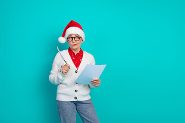 A cheerful elderly woman in a Santa hat and glasses holds a knife and a sheet of paper against a...