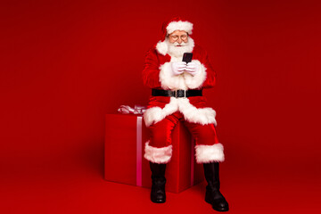 Santa Claus sits on a red gift box and shops on his phone in a Christmas themed studio with a bold red background
