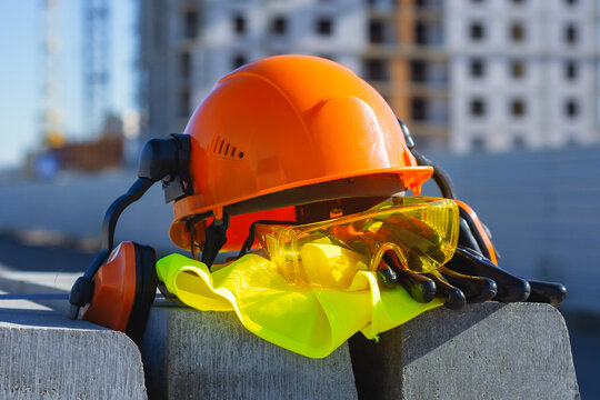 Construction safety equipment including a hard hat, protective gloves with goggles and ear defenders lies on a construction site. Concept workplace safety, industrial protection and gear. - Powered by Adobe