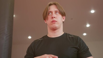 Athlete in black sportswear standing under bright ceiling lights with determined expression, preparing mentally for training session, showing focus, resilience inside professional gym environment