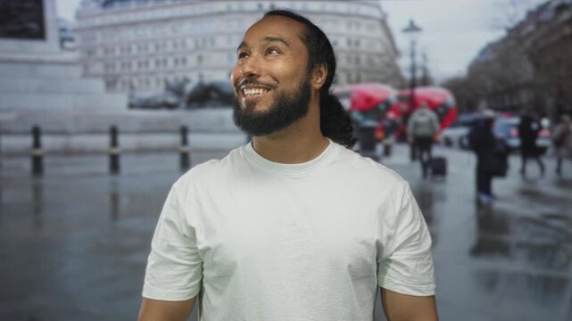 Bearded man in white t shirt smiling and looking up on wet street with red doubledecker bus behind him; optimism hope adventure.