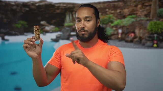 Man holding granola bar points finger to bar at poolside resort hotel deck outdoors while posing; wellness serenity.