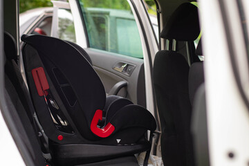 A close-up shot of a black child car seat with bright red latch elements, installed on the rear seat of a white car.