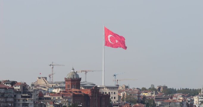Big Turkish national flag gently waving in wind with Istanbul cityscape in foreground. Long telephoto slow motion shot on hazy day, with Phanar Greek Orthodox College visible among urban buildings.