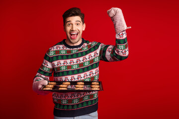 Funny christmas man in sweater bakes cookies on a tray wearing mittens in a bright red studio full of holiday joy and seasonal cheer
