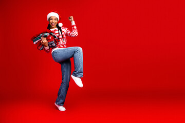 Fototapeta premium Funny cheerful young woman in santa hat jumps with gifts in a bright red studio creating christmas fun and holiday magic