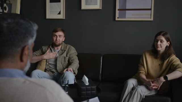 Over shoulder shot of male therapist facing couple sharing relationship problems with emotional man speaking while sitting on couch during consultation in therapists office