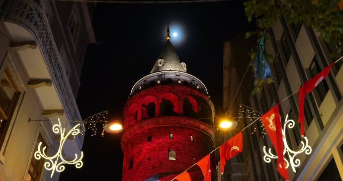 Red-illuminated Galata Tower with Turkish flag projection and full moon rising in background, low-angle shot. Red buntings hang across street, decorations celebrating 100th anniversary of republic - Powered by Adobe