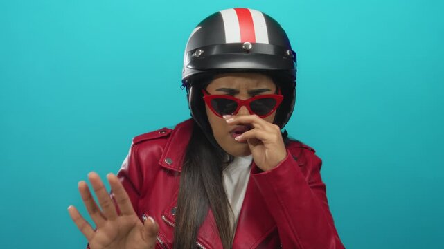 Woman holds nose to pinch foul air near a wall while wearing young hispanic helmet and standing by motorbike against a green isolated backdrop.
