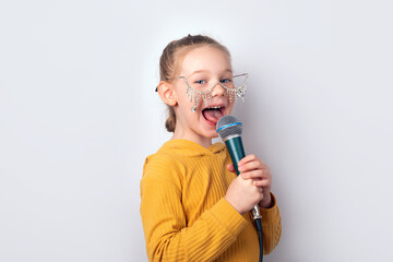 Cheerful young girl in yellow hoodie and playful glasses singing into a microphone against a plain white background.