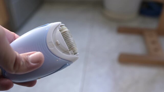 Close-up of an electric epilator turning on with buzzing motor sound, showing rotating tweezers and hand holding the device in soft daylight