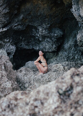 Young woman in black bikini sitting on rocks by sea cave