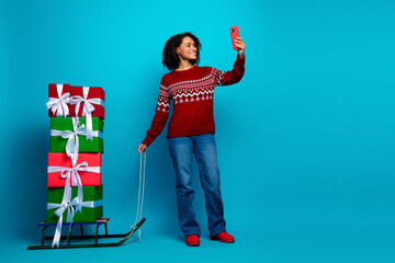 Funny holiday moment as a young woman takes a selfie beside a tall stack of wrapped gifts on a sled...