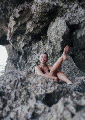 Smiling young woman in black bikini sitting on rocks at tropical beach