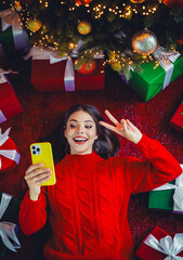 Playful holiday selfie under a twinkling Christmas tree with gifts and a smiling woman in a red...