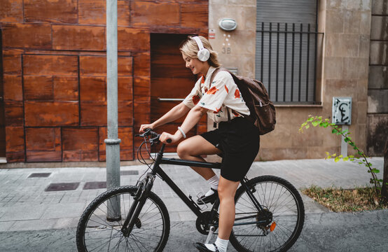 Young Asian woman pedals bike down narrow urban street, listening to music on headphones. Represents smart mobility, digital independence, and modern freelancing with wearable tech.