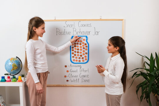 Two smiling schoolgirls participate in a classroom reward activity, adding stickers to a positive behavior jar. The concept promotes motivation, encouragement, teamwork, and learning success.