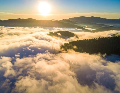 Aerial view of mountain range with clouds swirling below. The sun sets in the distance, casting a warm glow over the landscape