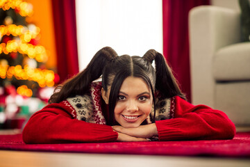 Joyful young woman in a cozy red sweater relaxing indoors with festive holiday decorations and a...