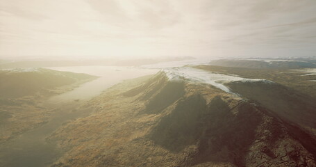 A vast mountainous terrain stretches under a hazy sky. Soft sunlight casts an ethereal glow over the peaks, revealing a serene lake amidst the rugged surroundings.