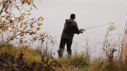Fisherman spinning for predatory fish on autumn lake, man in jacket casting lure from shore surrounded by reeds and fall foliage, active outdoor recreation and fishing adventure in nature