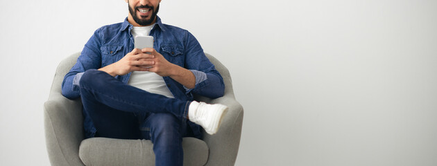 A man is seated in a cozy armchair, smiling as he uses his smartphone. He is dressed casually in jeans and a denim jacket, creating a relaxed atmosphere at home.