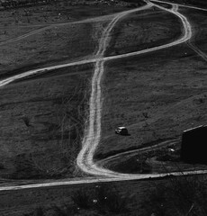 Black and white landscape with white SUV driving alone on curved dirt road across dry terrain. Minimal travel and solitude concept.