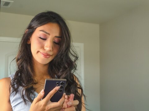 Latina using her phone and smiling in front a mirror - Powered by Adobe