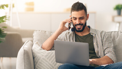 A man sits comfortably on a sofa, engaged in a phone conversation while typing on his laptop. The...