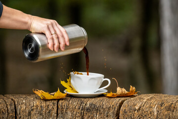 pouring hot coffee into a white ceramic cup outdoors on wooden surface surrounded by autumn leaves