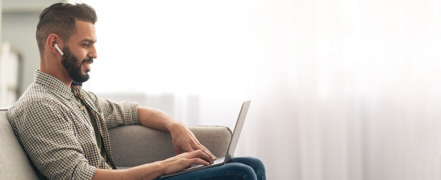 A man sits comfortably on a light-colored sofa, focused on his laptop while wearing earbuds. Soft natural light fills the room, creating a relaxing atmosphere for productivity.