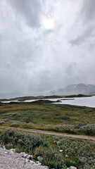 Gravel road along a mountain lake under cloudy sky. Scenic view of calm water, green slopes, and distant misty hills.