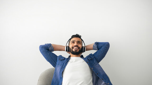 A young man smiles with joy as he listens to music using headphones while reclining on a soft chair. The serene setting emphasizes his relaxed mood in a bright indoor space.