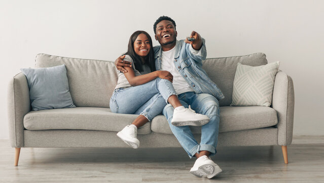 Beautiful African American black couple relaxing on sofa at home in living room, smiling man and woman spending free time together, watching TV or movie, embracing, laughing guy holding remote control