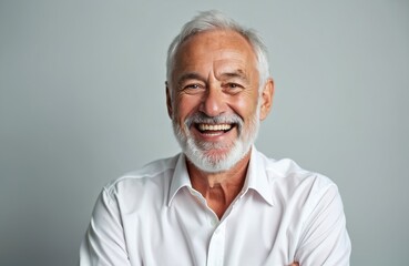 Smiling senior man with grey beard and hair wearing white shirt. Elderly male with toothy smile looking at camera on grey background. Happy mature person with confident expression.