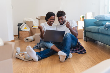 Joyful couple sitting among moving boxes, celebrating a successful online purchase or home setup while using a laptop during their move into a new home.