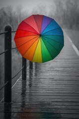A vibrant, multi-colored umbrella stands open against a moody, rain-swept backdrop, its bright hues a striking contrast to the muted tones of a wet wooden pier. The scene evokes a sense of resilience,