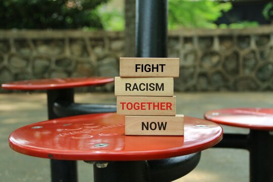 Bold wooden blocks spell out 'FIGHT RACISM TOGETHER NOW' on red park tables, promoting unity, inclusivity, and collective action against racism in a natural outdoor setting.