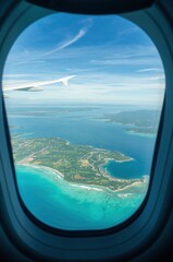 Viewed through an airplane window, a vibrant turquoise ocean meets lush green land dotted with buildings and fields, under a bright blue sky with wispy clouds. The sleek wing of the aircraft juts into