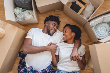 Happy African American couple lying on the floor surrounded by moving boxes, holding hands and smiling lovingly, celebrating new beginnings and the joy of moving into their first home together