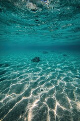 An immersive underwater perspective reveals a pristine sandy seabed illuminated by captivating patterns of sunlight filtering through the clear, tranquil turquoise water. Scattered rocks dot the seren