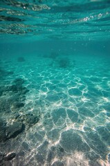 An inviting underwater view captures the serene beauty of a sun-drenched marine environment. Crystal clear turquoise water reveals a tranquil sandy seabed dappled with dancing sunlight patterns, along