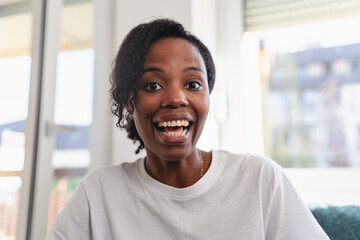 Portrait of a cheerful woman smiling brightly at home. Natural light, casual white shirt, and relaxed setting convey happiness, confidence, and positive energy. Having a video call. 
