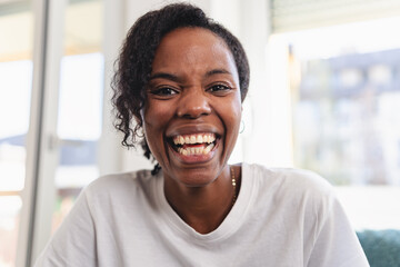Portrait of a cheerful woman smiling brightly at home. Natural light, casual white shirt, and relaxed setting convey happiness, confidence, and positive energy. Having a video call. 