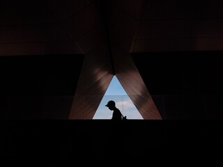 Silhouette of a person walking under geometric architecture with a triangular opening revealing blue sky, minimalist urban composition