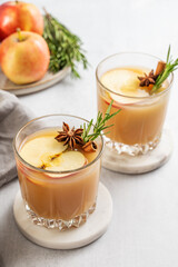 Two glasses of apple cider with cinnamon, anise, and rosemary on a light background with fresh fruits