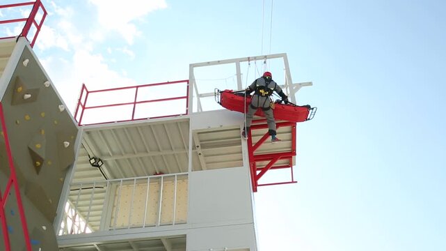 Rescue workers prepare for rappelling practice on a specialized high angle training tower, featuring a climbing wall against a bright sky, emphasizing readiness and skill development.