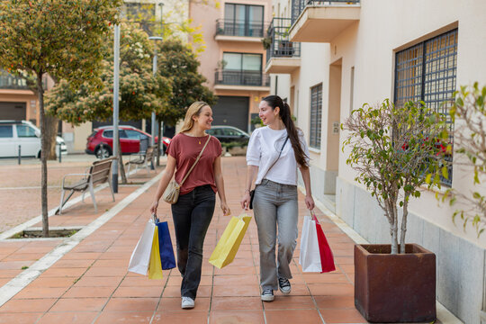 Happy female friends stroll through a residential street with shopping bags in hand, enjoying conversation and leisure time. Urban environment with trees and parked cars. Horizontal, wide angle.